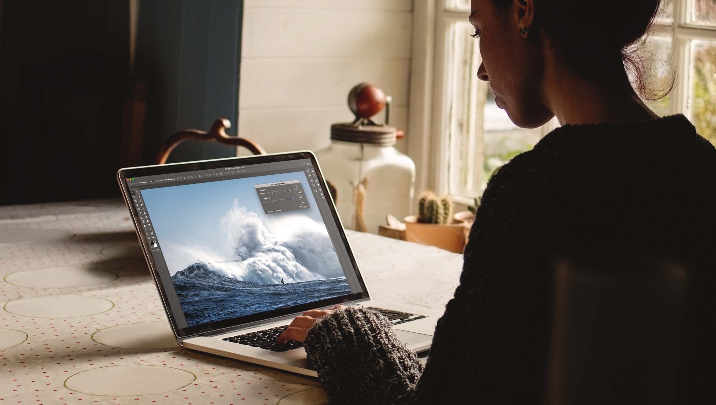 Woman sitting at kitchen table using Photoshop to edit image of surfer on large wave