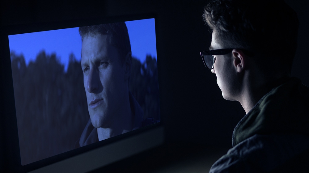 Teenaged boy looks at a television screen showing a man’s face. Teenaged boy looks at a television screen showing a man’s face.