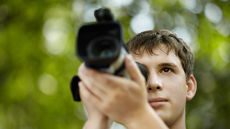 Young man holding video camera to his face stands in front of blurred out trees. Young man holding video camera to his face stands in front of blurred out trees.
