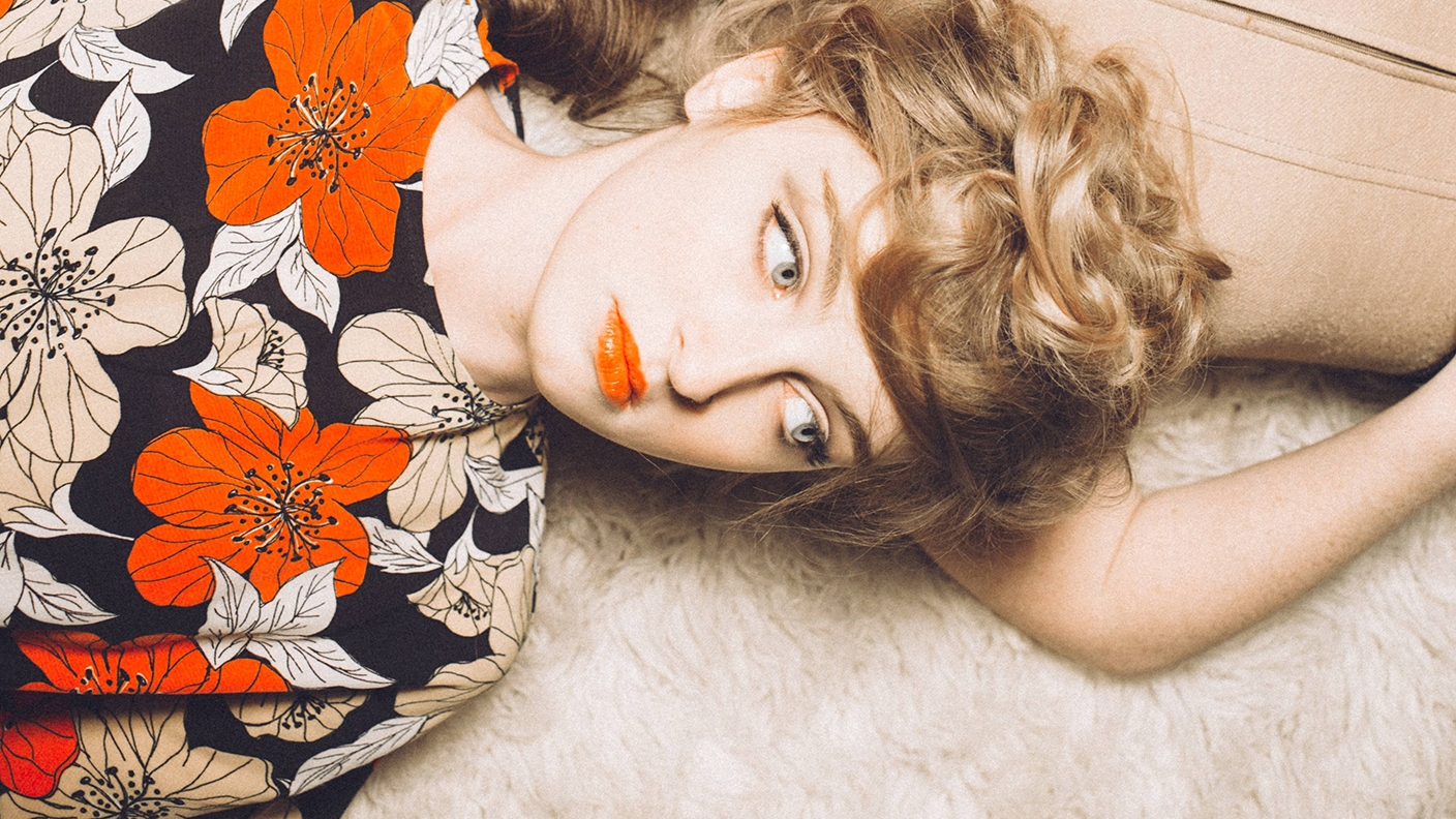 Woman lying on a rug with her head resting on a woman's shoulder. Woman is wearing a floral print top with matching lipstick.