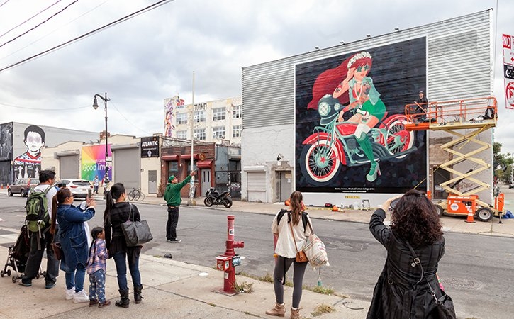 Des passants prennent en photo une fresque représentant une femme rousse vêtue de vert sur une moto rouge.