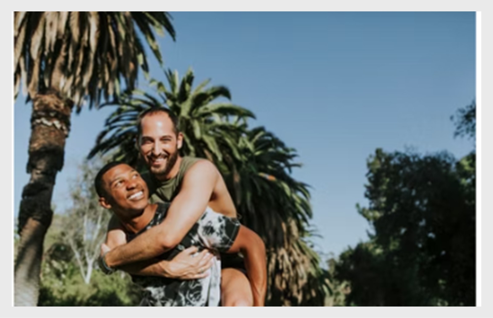 Foto av ett leende homosexuellt par utomhus med palmer i bakgrunden, där en person bär den andra på ryggen.